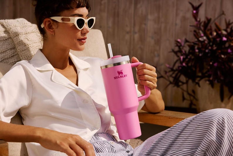 K&ouml;nnte beinhalten: Ein gro&szlig;er pinkfarbener Stanley-Becher mit Griff und Strohhalm. Der Becher hat einen wei&szlig;en Deckel und das Stanley-Logo. Die Person tr&auml;gt eine wei&szlig;e Sonnenbrille und ein wei&szlig;es Hemd.