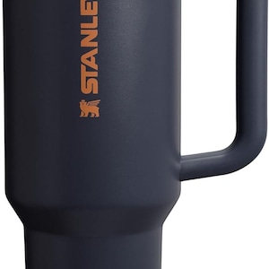 Puede incluir: Un termo de color azul marino con asa y tapa marr&oacute;n. El termo tiene la palabra "STANLEY" impresa en color cobre en el lateral. El termo est&aacute; dise&ntilde;ado para mantener las bebidas calientes o fr&iacute;as durante horas.