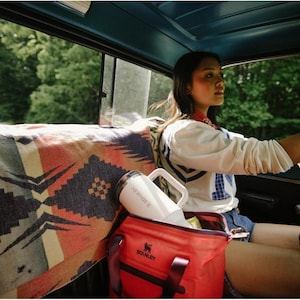 Puede incluir: Un termo Stanley blanco con asa y una bolsa nevera Stanley roja est&aacute;n dentro de un coche. Una manta estampada cubre el asiento trasero. La escena est&aacute; ambientada al aire libre, con &aacute;rboles visibles por la ventana.