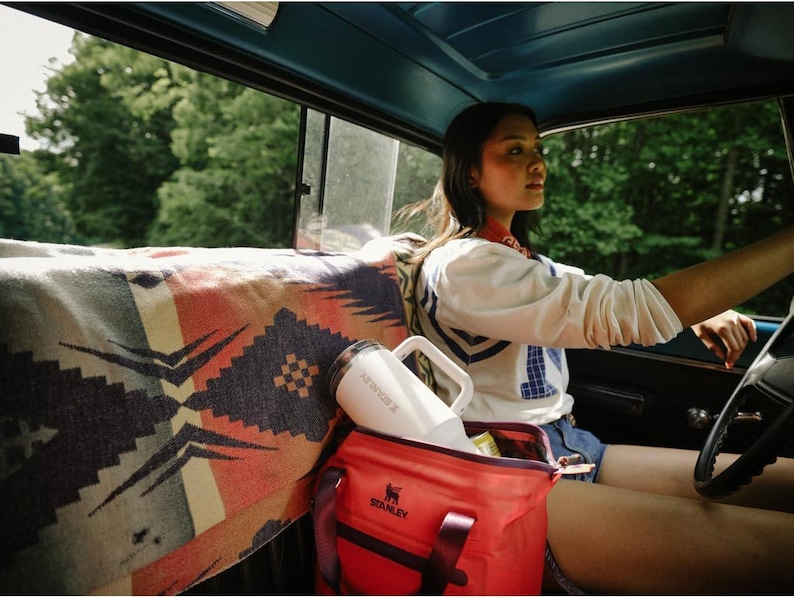 Puede incluir: Un termo Stanley blanco con asa y una bolsa nevera Stanley roja est&aacute;n dentro de un coche. Una manta estampada cubre el asiento trasero. La escena est&aacute; ambientada al aire libre, con &aacute;rboles visibles por la ventana.