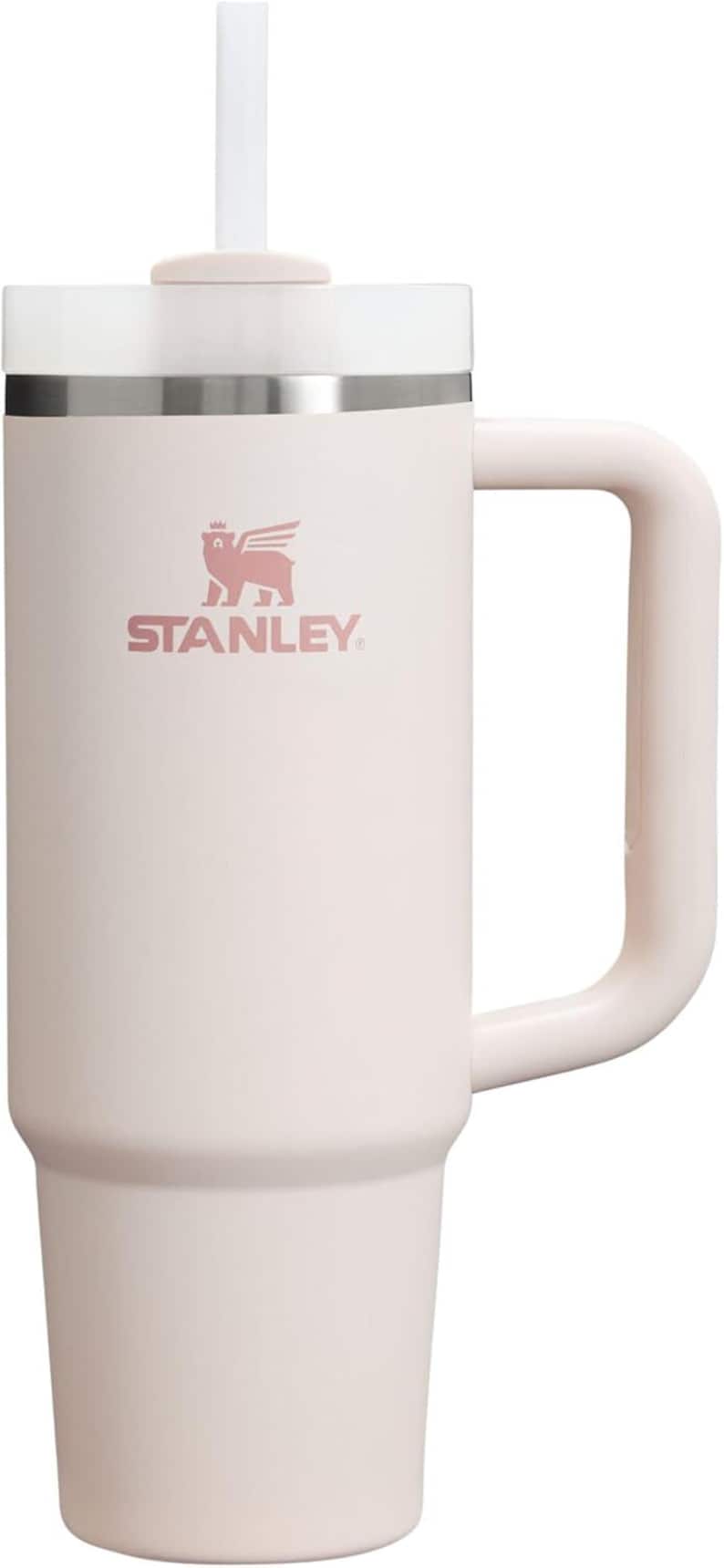 May include: A light pink Stanley Quencher H2.0 FlowState stainless steel tumbler with a handle and straw. The Stanley logo is printed on the front. The tumbler is designed to keep drinks cold for hours.