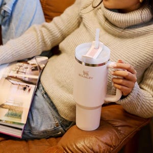 K&ouml;nnte beinhalten: Ein hellrosa Stanley-Becher mit wei&szlig;em Deckel und Griff. Der Becher hat einen Strohhalm und das Stanley-Logo. Der Becher steht auf einem braunen Ledersofa. Die Person tr&auml;gt einen grauen Pullover und blaue Jeans.