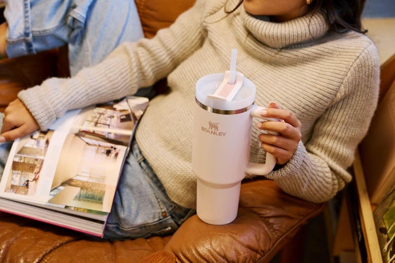 K&ouml;nnte beinhalten: Ein hellrosa Stanley-Becher mit wei&szlig;em Deckel und Griff. Der Becher hat einen Strohhalm und das Stanley-Logo. Der Becher steht auf einem braunen Ledersofa. Die Person tr&auml;gt einen grauen Pullover und blaue Jeans.