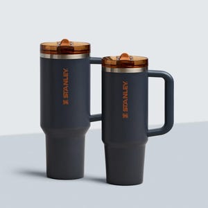 Puede incluir: Dos tazas de viaje Stanley de color gris oscuro con tapas y asas de color cobre. El logotipo de Stanley est&aacute; impreso en naranja en el lateral de cada taza. Una taza es m&aacute;s alta que la otra.