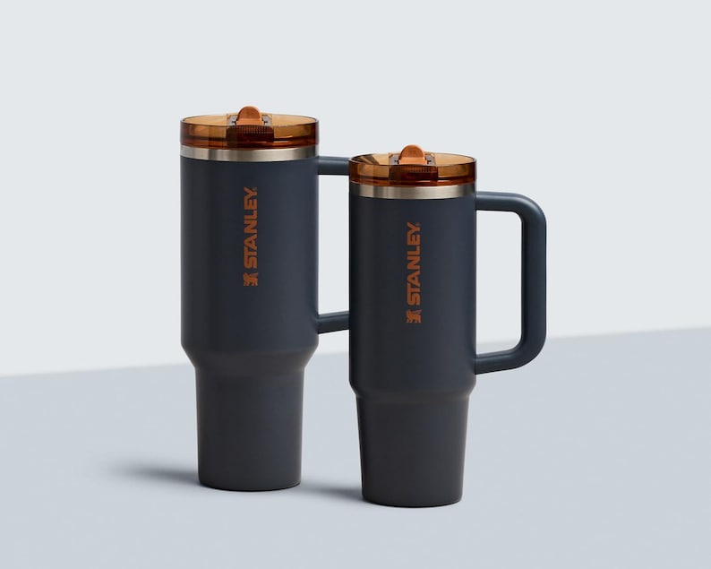 Puede incluir: Dos tazas de viaje Stanley de color gris oscuro con tapas y asas de color cobre. El logotipo de Stanley est&aacute; impreso en naranja en el lateral de cada taza. Una taza es m&aacute;s alta que la otra.