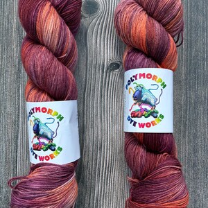 May include: Two skeins of hand-dyed yarn in shades of burgundy, orange, and peach. Each skein has a white label with the text "POLYMORPH DYE WORKS" and a cartoon sheep illustration. The yarn is arranged on a wooden surface.