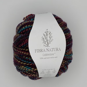 May include: A ball of Fibra Natura Cobblestone 100% superwash merino wool yarn in a variegated colorway of deep purple, teal, and gold. The white paper label features a botanical illustration and states 7 oz / 200 g / 612 yds / 560 m.