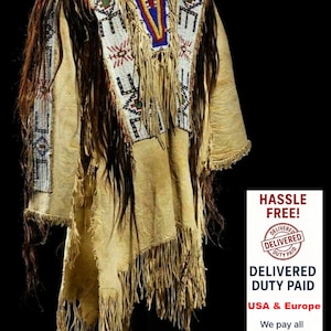 Puede incluir: Una prenda nativa americana de color beige con intrincados abalorios en blanco, rojo, azul y verde. La prenda presenta largas flecos y detalles de cabello castaño oscuro, mostrando la artesanía tradicional. La prenda se exhibe sobre un fondo negro.