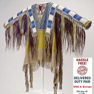 May include: A Native American-style garment with a V-neck, featuring a color palette of blue, yellow, and tan. The shoulders are adorned with white and blue beaded squares. Long fringe hangs from the sleeves and hem. The garment is displayed on a white stand.