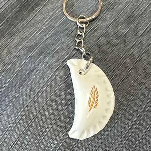 Könnte beinhalten: Weißer Keramik-Schlüsselanhänger in Halbmondform mit einem goldenen Weizen-Design. Der Schlüsselanhänger ist an einem silbernen Metallring und einer Kette befestigt.