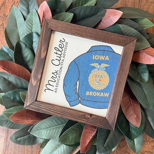 May include: A framed print featuring a blue FFA jacket design with the state of Iowa and the name Brokaw. The print is in a brown wooden frame and rests on a bed of green and brown leaves. Text on the print reads "Mrs. Cutler AG Education/FFA Advisor."