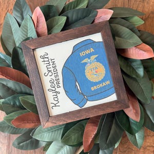 May include: Framed artwork featuring a blue FFA jacket design with the text "IOWA" and "BROKAW." The artwork also includes the name "Kaylee Smith" and the title "PRESIDENT." The frame is dark brown, and the artwork is set against a backdrop of green and reddish-brown leaves.