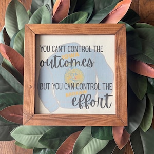 May include: A square wooden frame containing a white canvas with a light blue sweatshirt graphic. The text reads: YOU CAN'T CONTROL THE OUTCOMES BUT YOU CAN CONTROL THE EFFORT. The graphic includes an Iowa FFA emblem in yellow and blue.