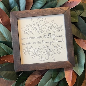 May include: A framed canvas sign with a floral design and the text "never underestimate the difference you make and the lives you touch".