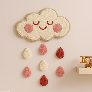 May include: A whimsical cloud-shaped wall hanging with a smiling face and rosy cheeks, accompanied by colorful raindrop accents. The cloud is cream-colored with a tan border, and the raindrops are in shades of pink and red. A charming nursery decoration.