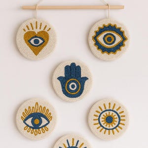 May include: A set of six round wall hangings with eye and hand designs. Each piece features a cream-colored background with blue and gold accents. The hangings are attached to a wooden dowel with a cream-colored string.