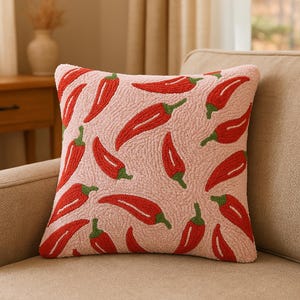 May include: A decorative square pillow with a light pink background and a pattern of red chili peppers with green stems. The pillow has a textured, tufted appearance and is placed on a beige sofa.