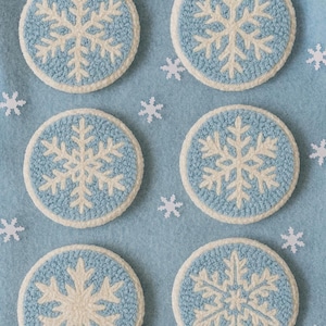 Könnte beinhalten: Sechs runde Untersetzer mit einem Schneeflocken-Design. Jeder Untersetzer hat eine hellblaue Mitte mit einer weißen Schneeflocke und einem weißen Rand. Die Untersetzer sind auf einer hellblauen Oberfläche angeordnet.