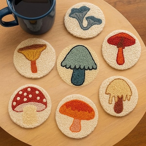 May include: A collection of seven round, off-white coasters with embroidered mushroom designs in various colors, including red, orange, yellow, and blue. A dark blue mug filled with a dark liquid sits in the upper left corner. The coasters are arranged on a wooden surface.