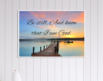 May include: A framed print featuring a serene lake scene at sunset. A wooden pier extends into the calm water, with a small boat in the distance. The sky is a blend of orange, blue, and yellow, with the text "Be still, And know that I am God".