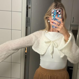 以下が含まれることがあります： 白いニットのクロップドカーディガン。長袖で、フロントにボウタイのディテールがあります。カーディガンは白いトップスと茶色のスカートの上に重ねて着ています。