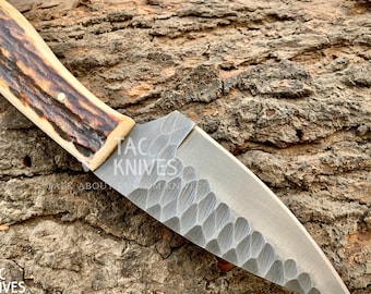 May include: A handcrafted knife with a textured blade and a handle made of light brown and dark brown material. The knife has a pointed tip and is set against a textured wooden surface. The text "TAC KNIVES" is visible.