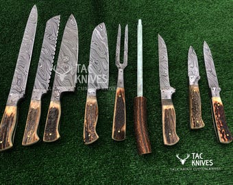 May include: A collection of custom knives with patterned blades and antler handles. The set includes various knife styles, a carving fork, and a sharpening steel. The blades have a unique, textured pattern. The knives are arranged on a green surface.