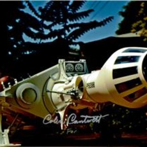 Puede incluir: Un modelo blanco de una nave espacial con un motor grande y redondo y una cabina con dos ventanas. El modelo está sentado sobre una superficie de madera con una firma que dice "Carl Cantrell" y "For."