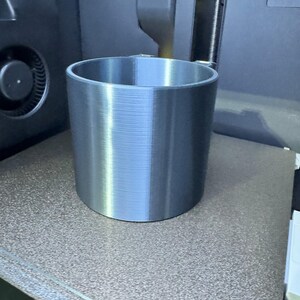 May include: A cylindrical, silver-colored object with a ridged texture, likely 3D-printed, sits on a speckled brown surface. The object is a cup or container, and the image is taken in a workshop setting, with machinery visible in the background.