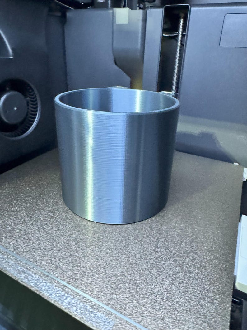 May include: A cylindrical, silver-colored object with a ridged texture, likely 3D-printed, sits on a speckled brown surface. The object is a cup or container, and the image is taken in a workshop setting, with machinery visible in the background.