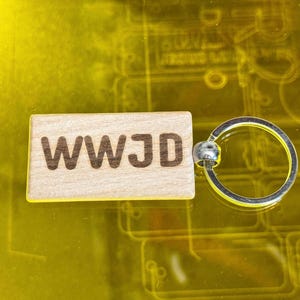 Pode incluir: Um porta-chaves retangular de madeira clara com o texto WWJD gravado em castanho escuro. A peça de madeira está ligada a um anel metálico circular de cor prateada, proporcionando uma forma funcional de prender chaves ou outros pequenos objetos.