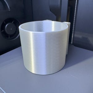 May include: A cylindrical, light-colored 3D-printed object with a handle, likely a cup or container. The object is made of layered material, showing the printing process. It sits on a gray surface, with a dark background.