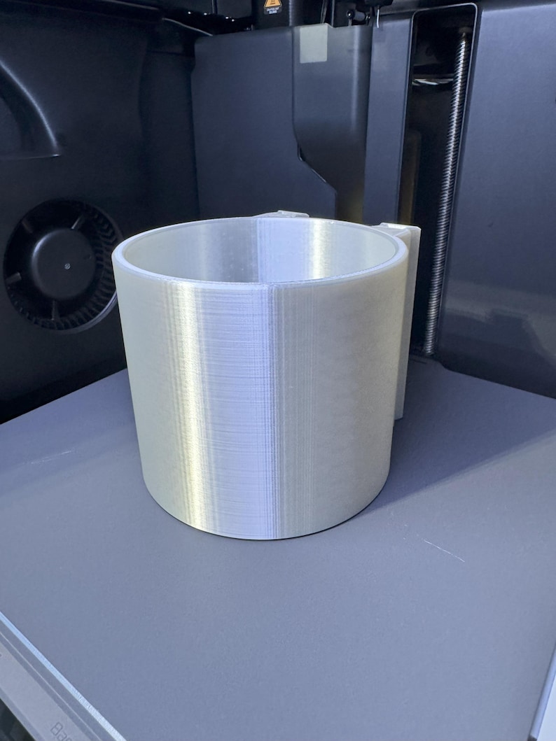 May include: A cylindrical, light-colored 3D-printed object with a handle, likely a cup or container. The object is made of layered material, showing the printing process. It sits on a gray surface, with a dark background.