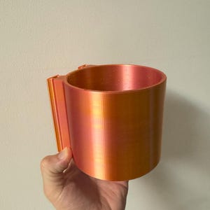 May include: A cylindrical, orange and red-toned cup with a handle. The cup is held by a hand against a plain white wall. The cup appears to be made of plastic or a similar material.