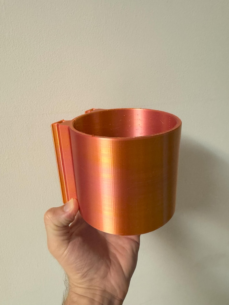 May include: A cylindrical, orange and red-toned cup with a handle. The cup is held by a hand against a plain white wall. The cup appears to be made of plastic or a similar material.
