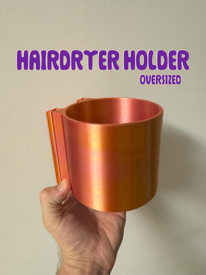 May include: An oversized, orange and pink hair dryer holder. The holder is cylindrical with a rectangular mounting bracket. The text "HAIRDRYER HOLDER OVERSIZED" is in purple above the holder.
