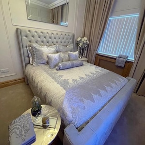 May include: A luxurious bed with a tufted headboard and matching upholstered frame. The bed is covered with a patterned comforter and several decorative pillows in shades of gray and white. A small side table holds a glass cloche and a mirrored tray.