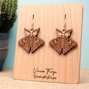 May include: A pair of wooden moth earrings with intricate carved details. The earrings feature a moth design with a skull, stars, and crescent moons. The earrings are displayed on a wooden stand with the text "Vireon Forge Handcrafted Designs".