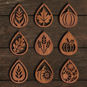 K&ouml;nnte beinhalten: Neun holzfarbene, tropfenf&ouml;rmige Ohrringe mit ausgeschnittenen Designs. Die Designs umfassen ein Blatt, ein Ahornblatt, einen K&uuml;rbis, ein Eichenblatt, Weizen, einen K&uuml;rbis mit Blumen, einen Zweig, einen K&uuml;rbis mit einer Sonnenblume und eine Sonnenblume.