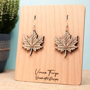 Puede incluir: Un par de pendientes de hoja de arce de madera marrón claro, exhibidos en un soporte de madera. Los pendientes tienen intrincados detalles de venas de hojas y ganchos plateados. El soporte tiene el texto "Vireon Forge Handcrafted Designs".