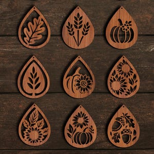 K&ouml;nnte beinhalten: Neun holzfarbene, tropfenf&ouml;rmige Ohrringe mit Herbstmotiven. Die Ohrringe zeigen ausgeschnittene Bilder von Bl&auml;ttern, Weizen, K&uuml;rbissen und Sonnenblumen. Die Ohrringe haben eine warme braune Farbe und werden auf einer dunkelbraunen Holzoberfl&auml;che pr&auml;sentiert.