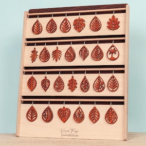 May include: A wooden display stand showcasing multiple pairs of leaf-shaped earrings. The earrings are made of reddish-brown wood and feature various intricate cut-out designs. The stand has a light wood frame with dark wood accents and the text "Vireon Forge Handcrafted Designs".