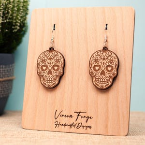 May include: A pair of wooden skull earrings with floral and heart details, hanging from silver hooks. The earrings are displayed on a light-colored wooden stand. The stand has the text "Vireon Forge Handcrafted Designs".