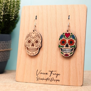 Può includere: Orecchini in legno a forma di teschio di zucchero. Un orecchino è non verniciato, l'altro è dipinto con dettagli rossi, arancioni, blu e verdi. Gli orecchini sono esposti su un supporto di legno con il testo "Vireon Forge Handcrafted Designs".