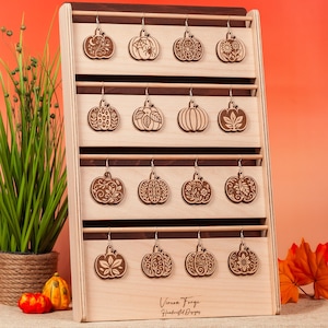 May include: Wooden pumpkin earrings displayed on a wooden rack. Each earring features a unique pumpkin design, including a spiderweb, floral patterns, and a sunflower. The rack is light-colored wood with a dark wood top. The text "Vireon Forge Handcrafted Designs" is visible.