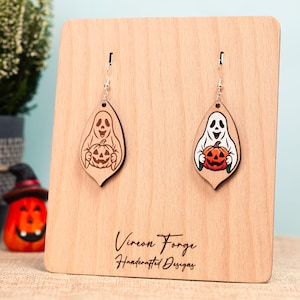 Peut inclure: Boucles d'oreilles d'Halloween en bois en forme de goutte d'eau. L'une est en bois naturel avec un fantôme sculpté tenant une citrouille. L'autre a un fantôme blanc tenant une citrouille colorée. Les boucles d'oreilles sont exposées sur un présentoir en bois.