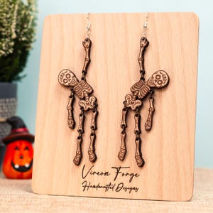 May include: Wooden skeleton earrings with intricate details, displayed on a wooden stand. The earrings feature a skull and articulated limbs. The stand has the text "Vireon Forge Handcrafted Designs". Perfect for Halloween or gothic-themed outfits.