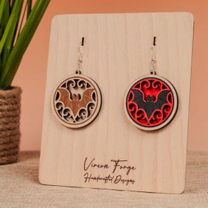 Può includere: Orecchini a pipistrello in legno esposti su un supporto in legno. Un orecchino presenta un disegno di pipistrello marrone all'interno di una cornice circolare marrone chiaro. L'altro orecchino ha un disegno di pipistrello nero con sfondo rosso e cornice circolare nera.