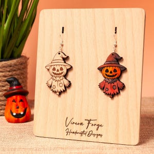 Könnte beinhalten: Holz-Halloween-Ohrringe mit einem Vogelscheuchen- und Kürbis-Design. Die Ohrringe werden auf einem Holzständer mit dem Text "Vireon Forge Handcrafted Designs" präsentiert. Der Vogelscheuchen-Ohrring ist hellbraun, der Kürbis-Ohrring ist orange und rot.