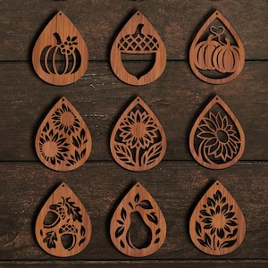 K&ouml;nnte beinhalten: Neun holzfarbene Ohrringe in Tropfenform mit ausgeschnittenen Designs. Die Designs umfassen K&uuml;rbisse, Eicheln, Sonnenblumen und eine Birne. Die Ohrringe haben eine warme braune Farbe und sind auf einer dunklen Holzoberfl&auml;che angeordnet.